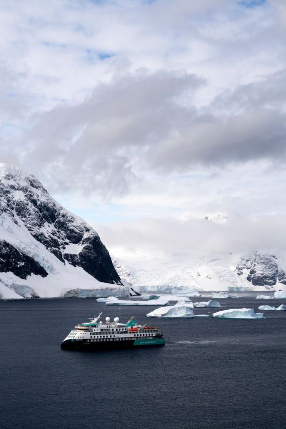 A cruise ship sails past icebergs and snowy mountains in Antarctica on a cloudy day.